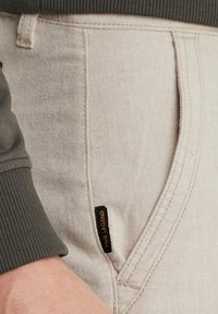 Pantalones de algodón beige con un acabado texturizado, que cuentan con un bolsillo lateral y una pequeña etiqueta negra que dice "PAINE LEGEND". Detalles en verde oliva.