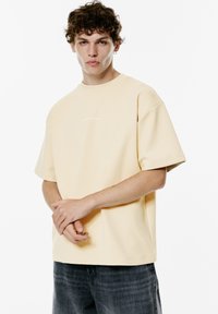 Lys gul bomulds t-shirt med korte ærmer, rund halsudskæring og et subtilt tekstdesign på forsiden. Modellen står med armene over kors.