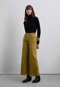 Mujer vistiendo un suéter de cuello alto negro, pantalones anchos mostaza con adornos metálicos y botines negros, posando contra un fondo gris liso.