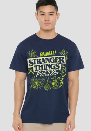 Man wearing navy blue Stranger Things T-shirt with neon yellow designs including text, bike, radio, and skull illustrations.