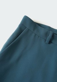 Tissu turquoise, lisse, avec une taille et un design de pli latéral, présentant un détail de fermeture cachée pour un aspect épuré.