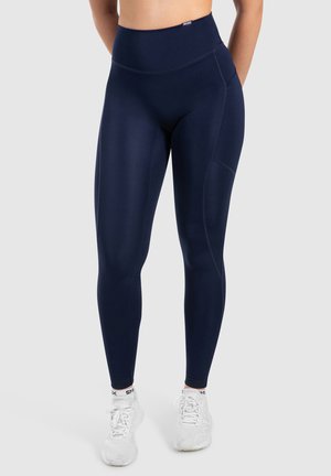 Marine hoge taille leggings van rekbaar materiaal met details aan de zijkanten. Gecombineerd met witte sportschoenen met gekleurde accenten.