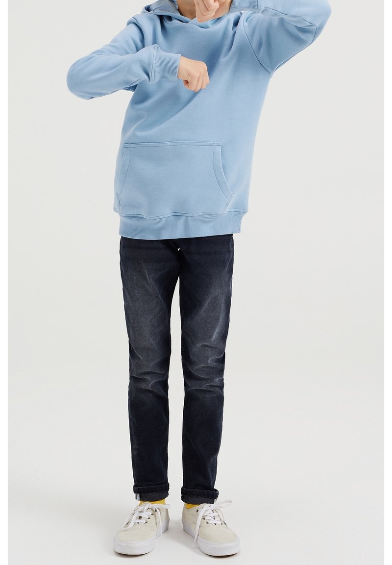 Světle modrá mikina s kapucí a klokaní kapsou, v kombinaci s tmavými džíny a bílými teniskami s žlutými ponožkami. Jednoduchý design.