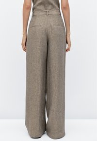 Achteraanzicht van een persoon in hooggelegde, wijde, lichtbruine linnen broeken met riemlussen en achterzakken.