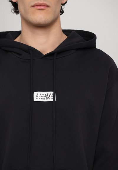 Haut à capuche noir avec une poche frontale, un cordon de serrage et une étiquette blanche rectangulaire avec des chiffres et un logo circulaire.