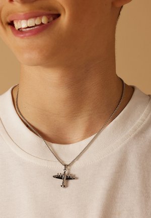Zilveren ketting met een hanger in de vorm van een vliegtuig, hangend aan een dunne ketting, gedragen over een wit sweatshirt.
