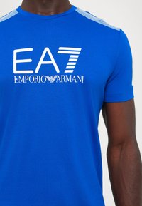 Blue cotton t-shirt featuring a white EA7 logo and "EMPORIO ARMANI" text. Horizontal white stripes accent the shoulders. Short sleeves.