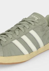 Ανοιχτοπράσινο sneaker Adidas με λευκές ρίγες, σχέδιο με κορδόνια και μπεζ υφή στη σόλα σε λευκό φόντο.