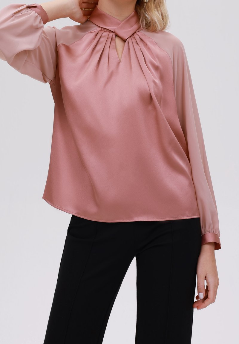 Blouse rose avec finition satinée, devant plissé, encolure à trou de serrure et manches longues transparentes. Assortie à un pantalon noir ajusté.