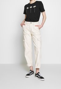 Camiseta negra con gráficos de margaritas, pantalones cargo crema con múltiples bolsillos y zapatillas Vans negras con acentos blancos.