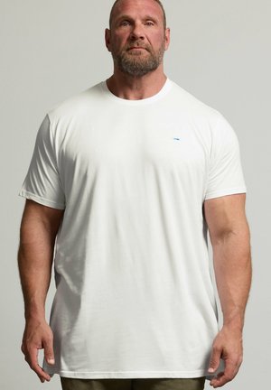 Umięśniony mężczyzna z brodą stoi przodem, ubrany w prosty biały t-shirt z krótkim rękawem i oliwkowe spodnie na neutralnym tle.