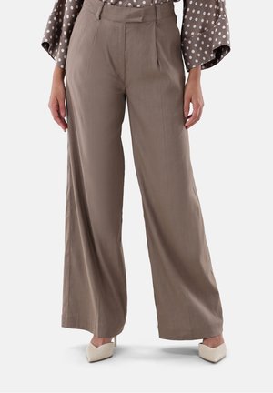 Vrouw draagt taupekleurige broek met hoge taille en wijde pijpen, met een bruine en witte polkadot blouse en witte puntige hakken, staat rechtop.