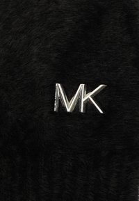 Tissu en fausse fourrure noire présentant un logo "MK" argenté brillant. La texture est douce et moelleuse, contrastant avec la quincaillerie métallique.