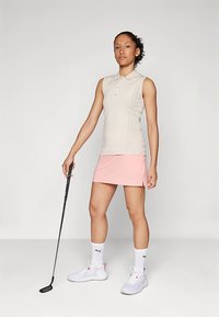 Világos barnás bézs ujjatlan galléros póló, rózsaszín golf szoknyával párosítva. Fehér sportcipő és zokni. Fekete golfütőt tart.