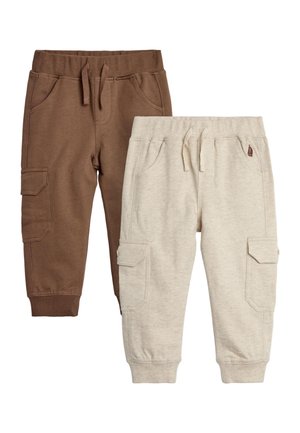 Peuter draagt een blauwe dinosaurus-hoodie met kleurrijke spikes, een bruine broek en sneakers, zit op een witte kubus tegen een roze achtergrond.