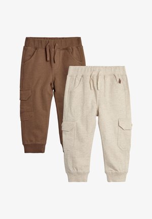 Peuter draagt een blauwe dinosaurus-hoodie met kleurrijke spikes, een bruine broek en sneakers, zit op een witte kubus tegen een roze achtergrond.