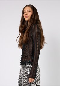 Schwarzes langärmeliges Hemd mit Spitzenstruktur, figurbetontem Design und transparenten Ärmeln, kombiniert mit gemusterten, hoch taillierten Hosen in monochromen Tönen.