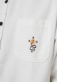 Camicia in tessuto a coste bianco con taschino ricamato con un serpente che si attorciglia attorno a un cerchio arancione, circondato dalle lettere K, A, O in filo nero.