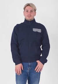 Giacca a zip blu navy con colletto alto, tasca sul petto grigia e polsini abbinati. Presenta un tessuto testurizzato e tasche frontali.