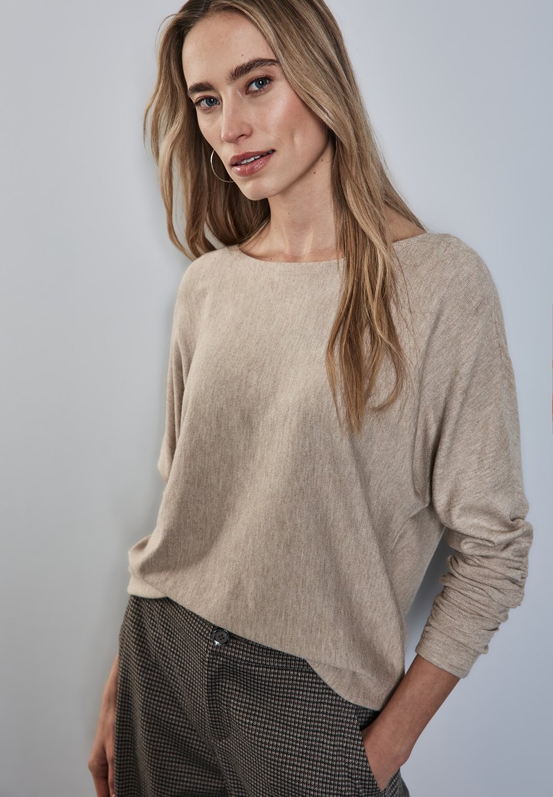 Maglione loose-fit beige chiaro realizzato in un tessuto a maglia testurizzato, abbinato a pantaloni grigi a quadri con dettaglio dei bottoni.
