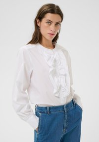 Jonge vrouw met lang bruin haar draagt een witte blouse met ruffles en blauwe hooggemonteerde jeans, staande met haar handen in haar zakken tegen een witte achtergrond.