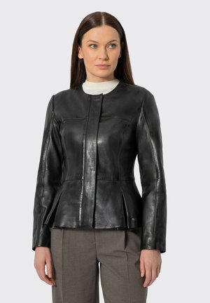 Žena v přiléhavé černé kožené bundě s rozparky, bílém topu s vysokým límcem a šedých kalhotách, stojící před jednobarevným pozadím.