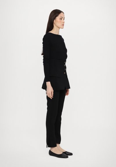 Fekete kötött felső hosszú ujjal és fitted derékkal, peplum fazonnal és dekoratív gombokkal. Slim-fit fekete nadrággal és lapos cipőkkel párosítva.