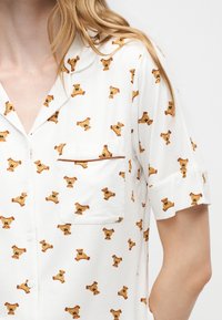 Chemise blanche à manches courtes avec motif ours en peluche marron et poche poitrine, portée par une personne aux longs cheveux blonds visibles.