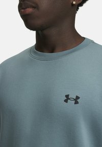 A close-up of a young person’s neck and shoulder, wearing a light blue sweatshirt featuring a black Under Armour logo.