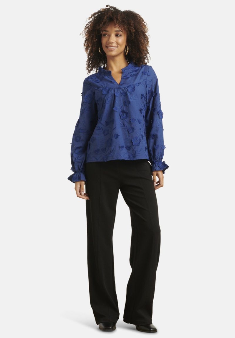 Blue floral embroidered blouse with ruffled collar and long sleeves, paired with black wide-leg trousers and black shoes.