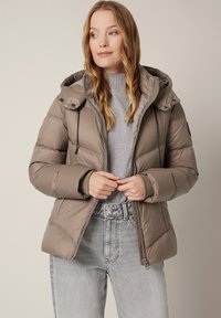 Kurtka puchowa w kolorze taupe z odpinanym kapturem, zamkiem błyskawicznym oraz czarnymi sznurkami. Noszona na szarej bluzie, w zestawie z lekko przetartymi dżinsami.