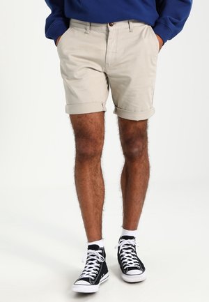 Personne portant un short beige retroussé, une chemise à manches longues bleue, et des baskets montantes noires avec des lacets blancs et des chaussettes blanches.
