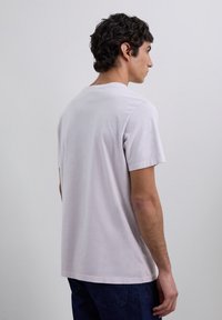Joven con cabello rizado oscuro que lleva una camiseta básica de manga corta gris claro y jeans oscursos, de perfil frente a un fondo claro.