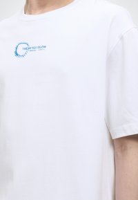 Λευκό κοντομάνικο μπλουζάκι με μπλε κυκλικό σχέδιο και κείμενο "THE AFTER GLOW" στο αριστερό στήθος, που φοριέται από άτομο.