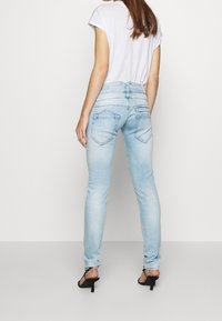 Hellblaue Skinny-Jeans mit tailliertem Design, ausgestattet mit zwei hinteren Taschen mit Stickakzenten und einer mittelhohen Taille. Kombiniert mit schwarzen Sandalen.