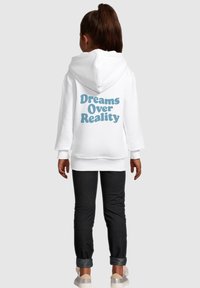 White hooded sweatshirt with blue text "Dreams Over Reality" on the back. Paired with dark pants and light shoes, facing away from the camera.