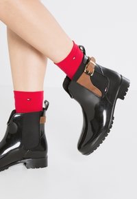 Des jambes portant des chaussettes rouges et des bottines noires brillantes avec des sangles en cuir marron, se tenant sur une surface blanche.