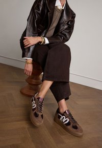 Braune Wildleder- und Ledersneaker mit metallischen Akzenten, rundem Zeh und dicker Gummisohle. Kombiniert mit dunklen Hosen und einer braunen Jacke.