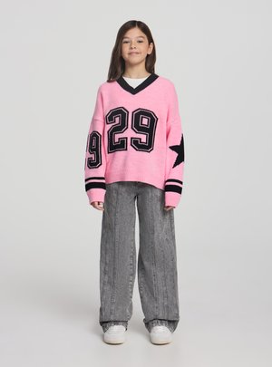 Ragazza che indossa un maglione rosa con il numero 29 nero e disegni di stelle, jeans grigi larghi e sneakers bianche, in piedi contro uno sfondo grigio.