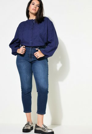 Camicia blu marina con bottone e motivo testurizzato, abbinata a jeans in denim scuro aderenti e mocassini metallici, che mostrano una silhouette rilassata.