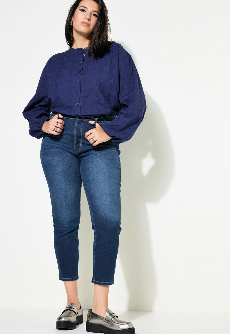 Marinblå blus med knappar och texturerat mönster, tillsammans med åtsittande mörka denimjeans och metalliska loafers, vilket ger en avslappnad silhuett.