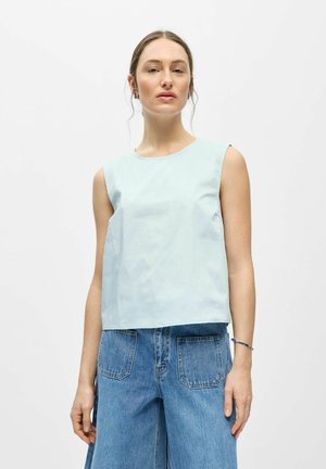 Femme portant un haut bleu clair sans manches et un jean bleu ample avec des poches avant, debout sur un fond blanc uni.