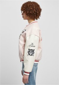 Lys rosa bomberjakke med hvite ermer og røde striper. Har broderte design, inkludert tekst og logoer på ermet.