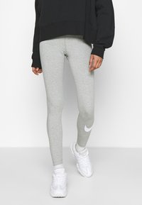Leggings atletici grigio chiaro realizzati in tessuto elasticizzato, con un logo Nike bianco sul polpaccio sinistro. Indossati con una felpa nera oversize.