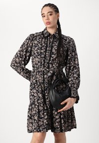 Abito a stampa floreale con maniche lunghe, sfondo nero e fiori dai colori chiari. Caratterizzato da una vita arricciata e una gonna a moretti. Porta una borsa nera.