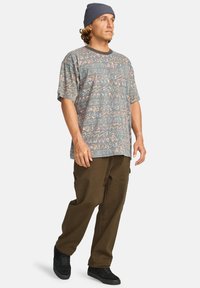 Camisa de manga corta gris con un patrón de diseños abstractos multicolores, combinada con pantalones sueltos marrones y zapatillas negras.