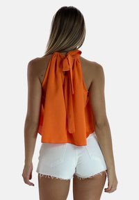 Top arancione con scollatura a halter e dettagli in fiocco sul retro, caratterizzato da una vestibilità ampia e tessuto leggero. Abbinato a pantaloncini di jeans bianchi con strappi.