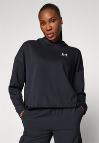 Sudadera corta negra hecha de una tela suave, con un bolsillo frontal y un pequeño logo blanco en el pecho. Usada con pantalones oscuros a juego.