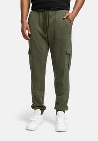 Man in olijfgroene cargo joggers en witte sneakers, staand met één hand in de zak tegen een effen achtergrond.