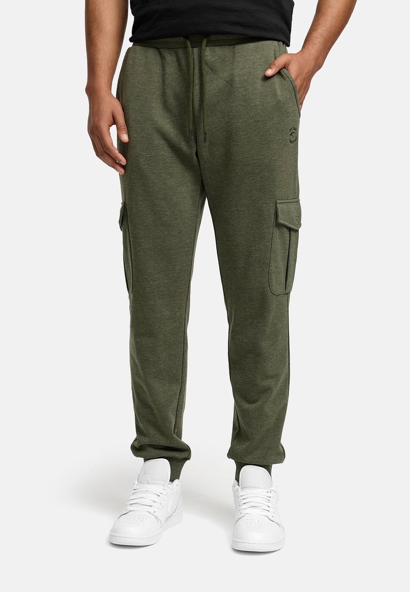 Man in olijfgroene cargo joggers en witte sneakers, staand met één hand in de zak tegen een effen achtergrond.